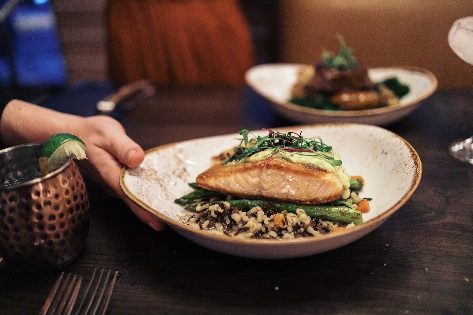 A person setting down a plate of cooked fish that is sitting on top of a bed of veggies and grain.