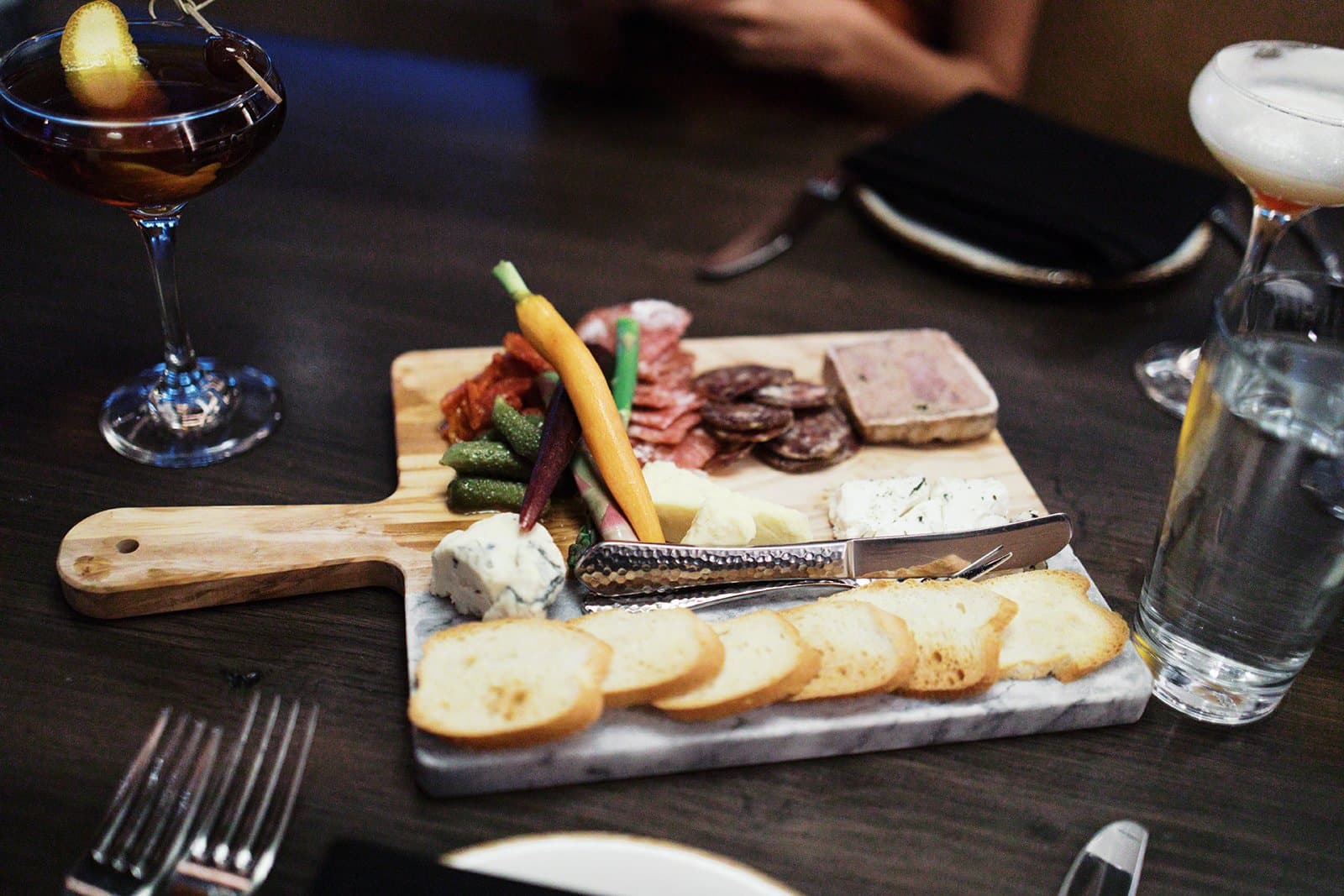 An array of mixed charcuterie meats and cheeses with pickled vegetables and crispy crackers on top of a half wooden and marble board.