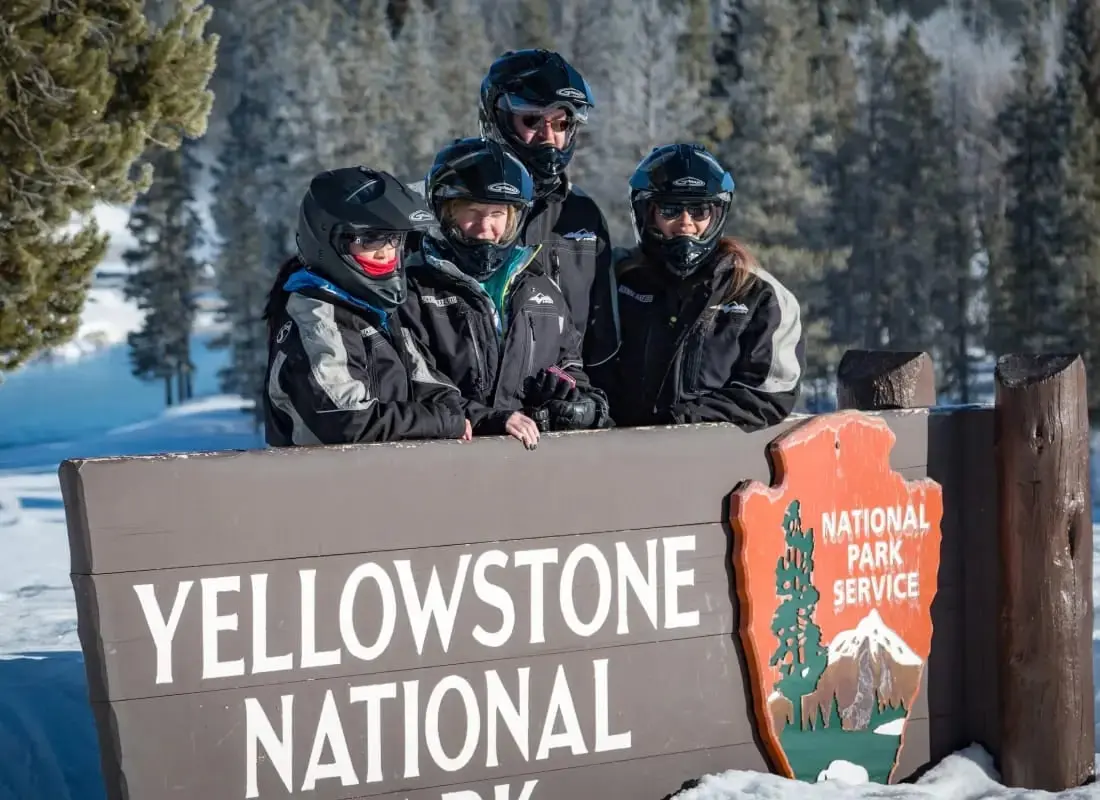 A photo of four people wearing snowmobile helmets pose behind a Yellowstone National Park sign