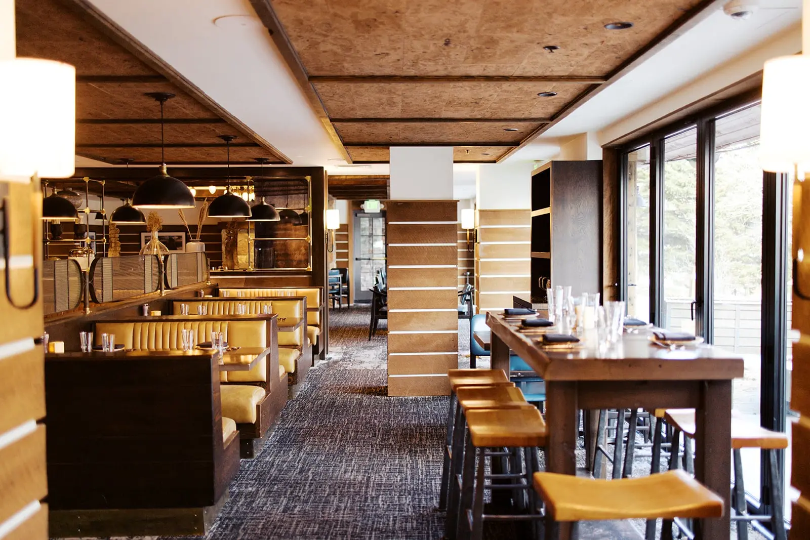 A photo of an aisle in a restaurant, on one side are U-shaped booths and the other side is a long communal wooden table with bar stools