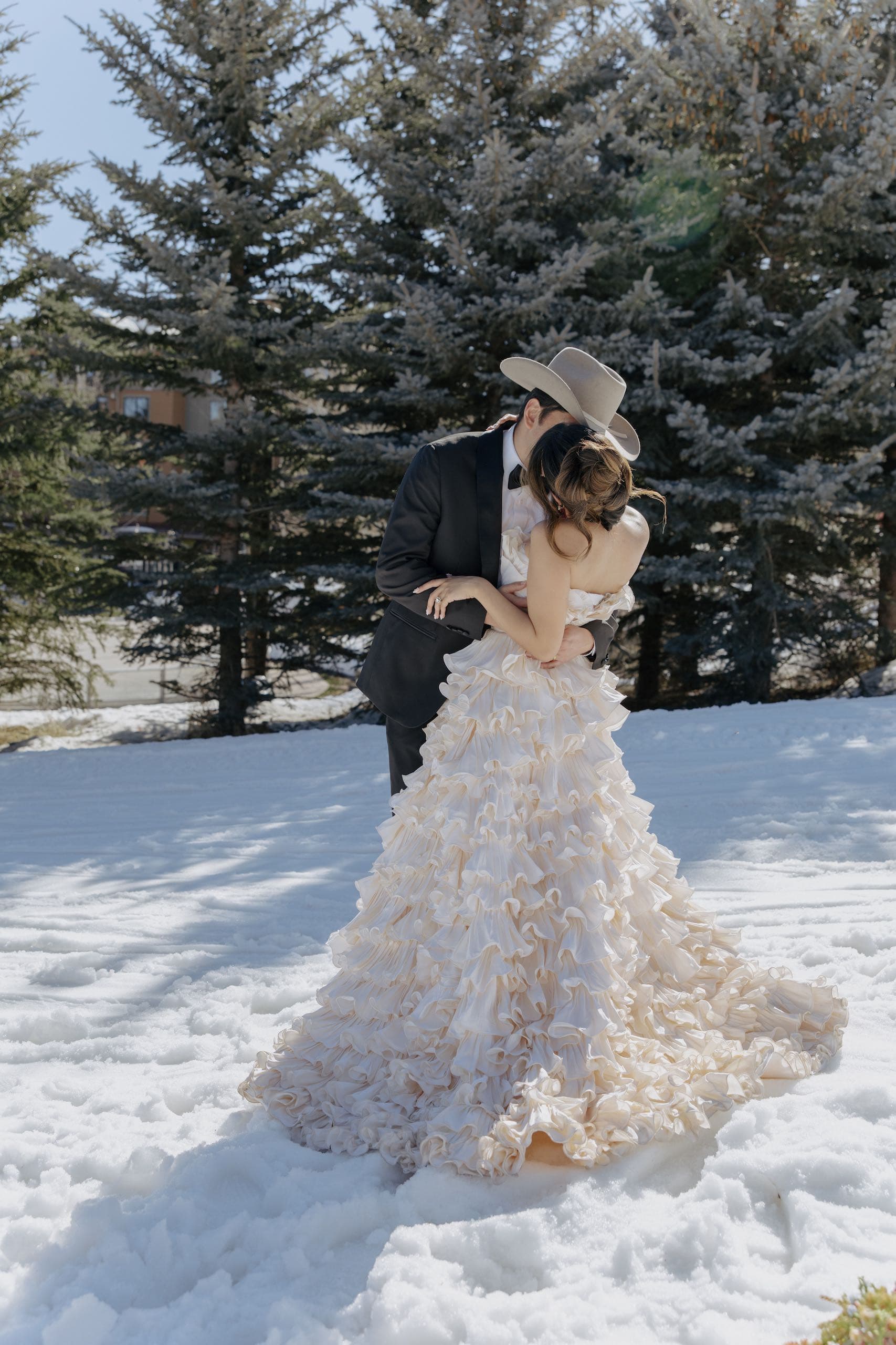 A photo of a wedded couple kissing in a snowy field. The man is wearing a light-coloured cowboy hat.