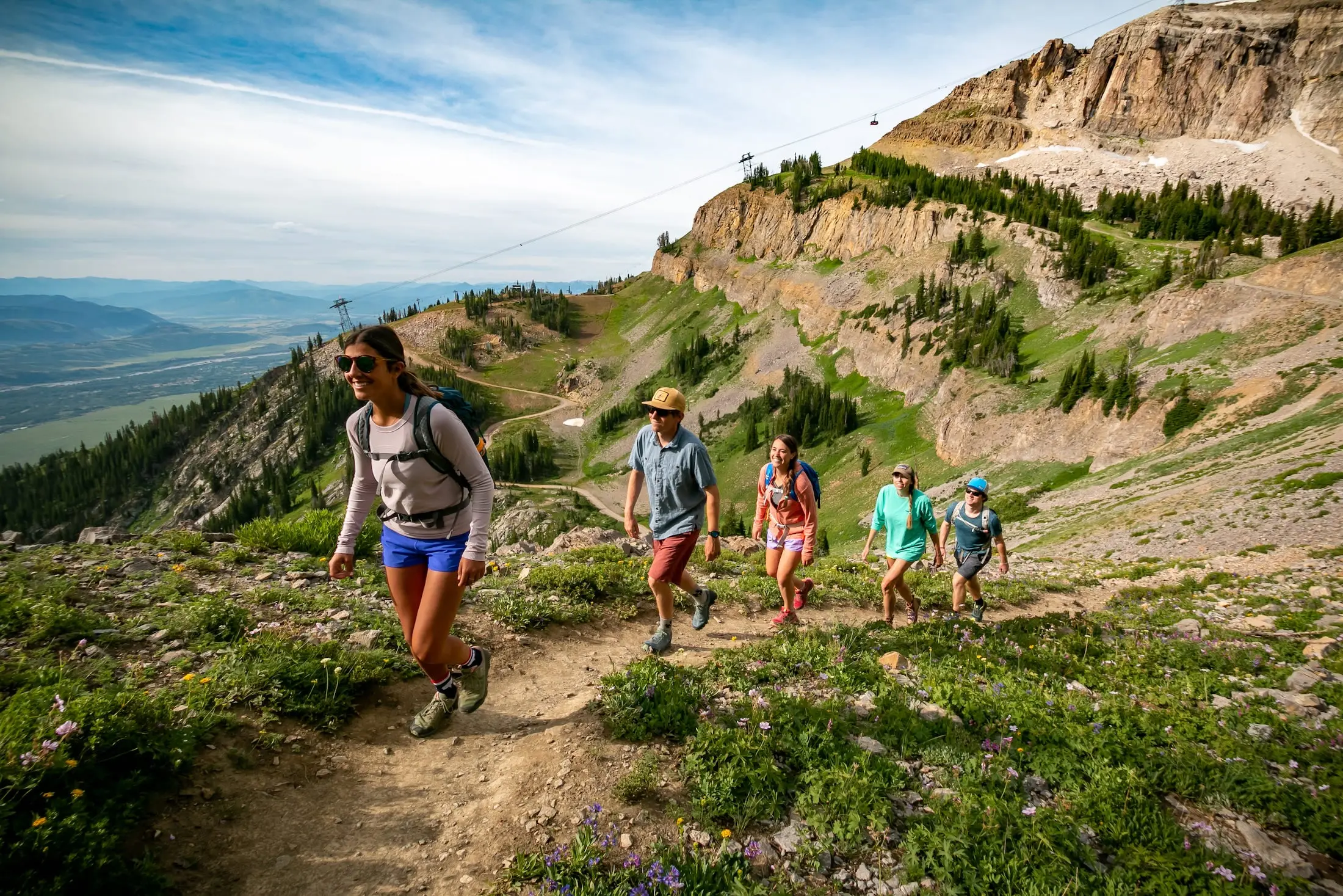 A group of five trail runners hike up through a mountainous plain with an aerial tram in the background