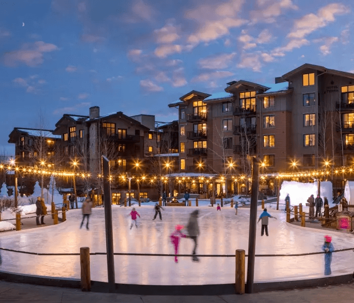 A group of people skate on a lit open-air fenced in ice rink during the blue hour