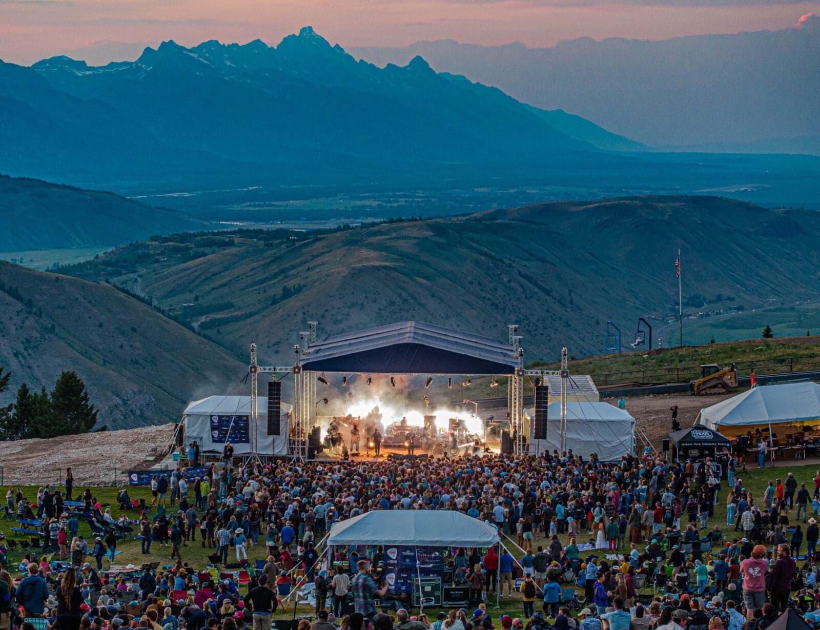 An aerial view of an outdoor concert with a covered and lit stage with a band playing and a massive group of people, backdropped by a vast mountain landscape during the blue hour
