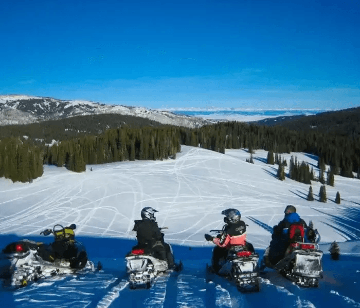 Three people in snowmobiling gear on snowmobiles on a snowy hill