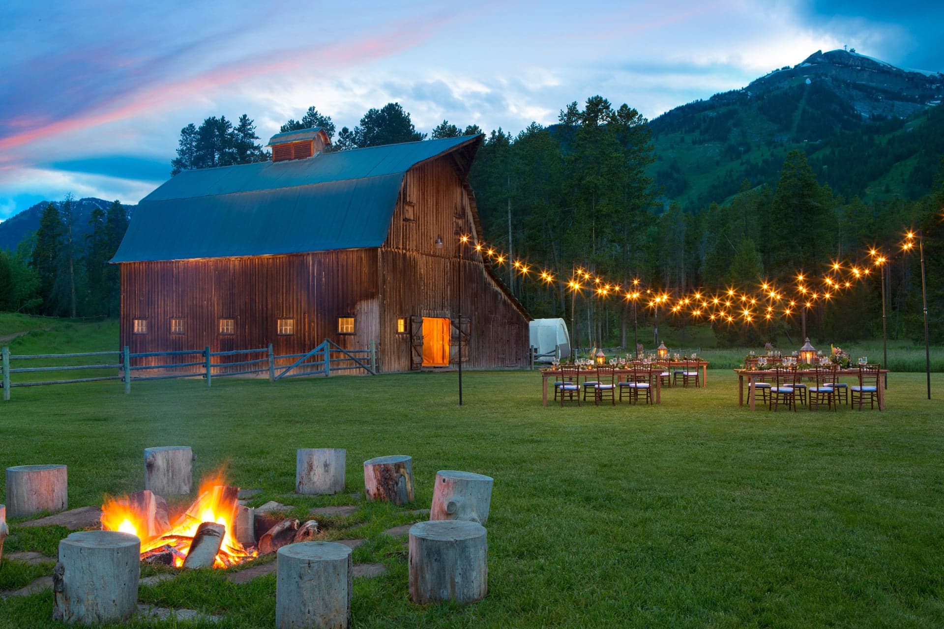 A rustic barn in the evening, with multiple tables with place settings. A fire with log stools can be seen in the foreground.