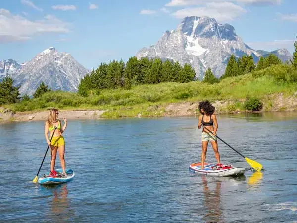 Two women stand-up paddle boarding on a lake amidst the backdrop of a mountain range